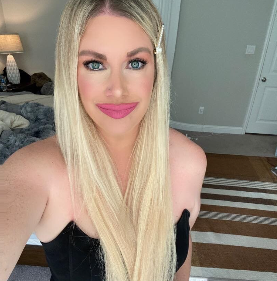 Osoba s dlhými blond vlasmi v interiéri, oblečená v čiernych šatách, s jemným líčením a ružovým rúžom.