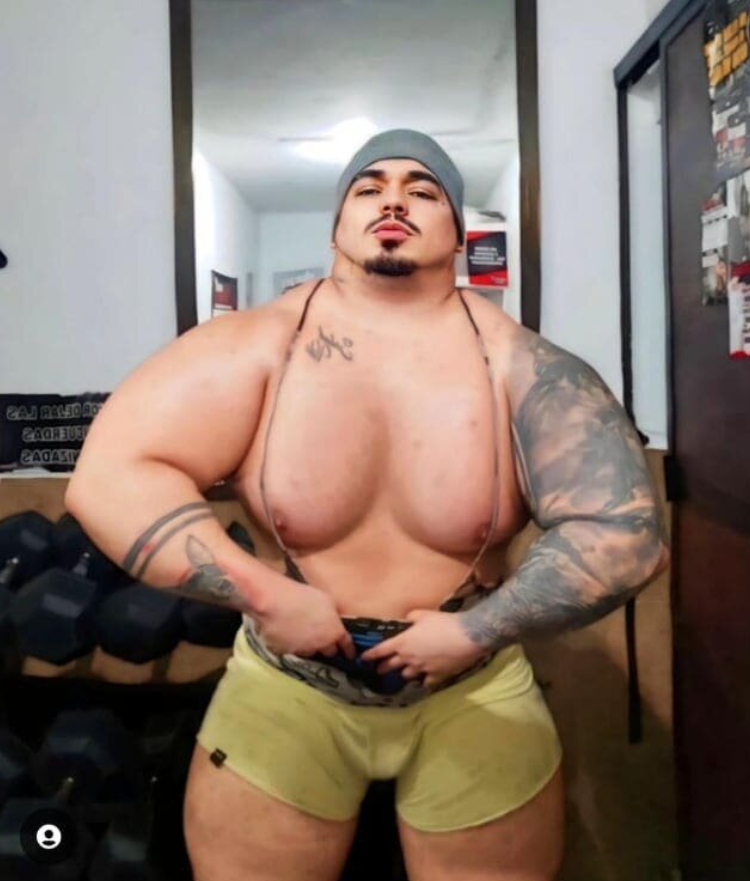 Osoba s výraznými svalmi a tetovaním stojí v telocvični s činkami na pozadí.