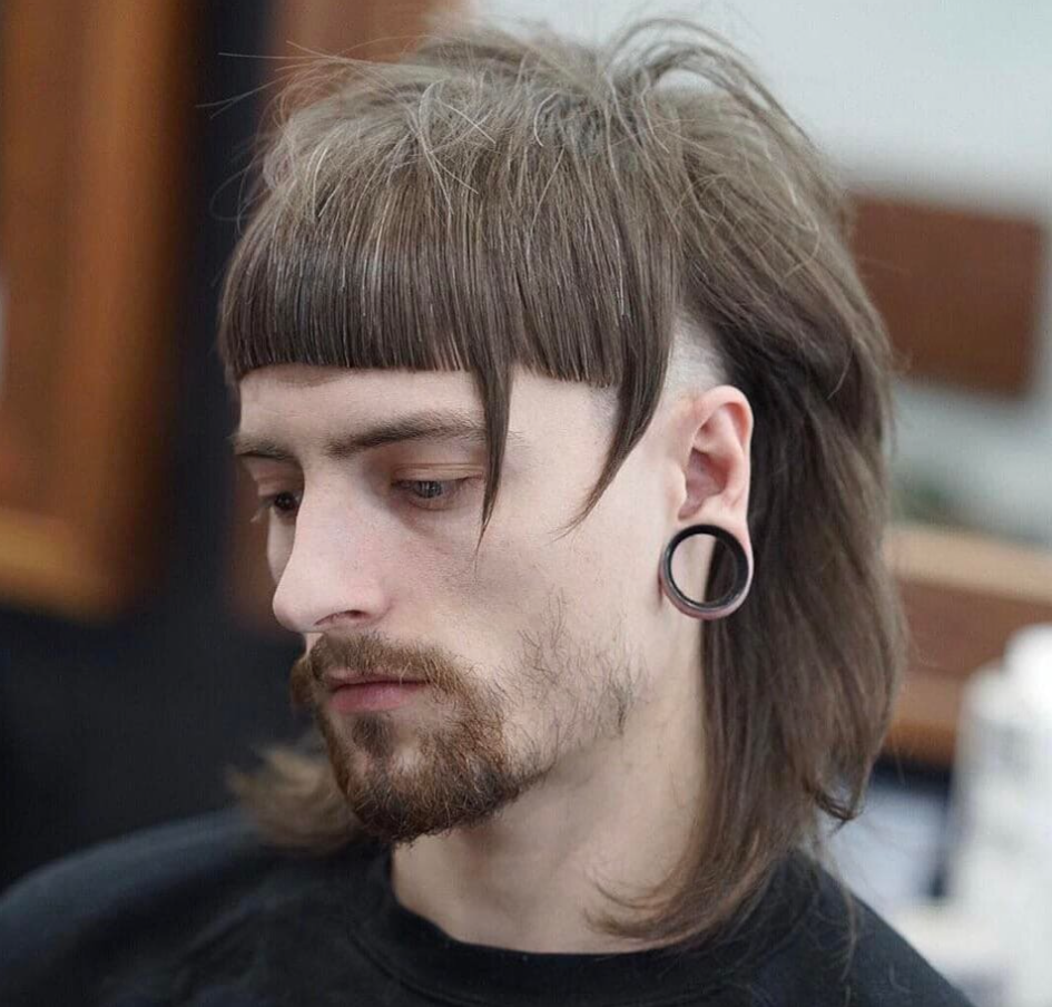 Ostrihaný muž s kruhovými náušnicami a dlhými vlasmi po stranách na moderný účes v holičstve.