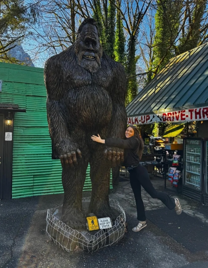 Žena objíma sochu veľkého Bigfoota pred zelenou budovou so stánkom a stromami v pozadí.