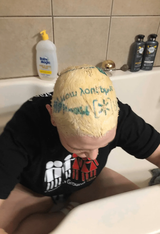 Osoba s krémom na hlave sedí v bielej vani; na stene sú vidieť produkty na kúpanie.