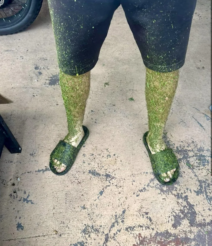 Mužské nohy pokryté trávou v šortkách a šľapkách po práci v záhrade, stojace na betónovom povrchu.