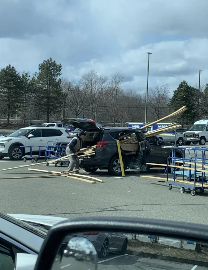 Auto preplnené drevom na parkovisku, drevo trčí z kufra a zadných dverí, osoba upravuje náklad.