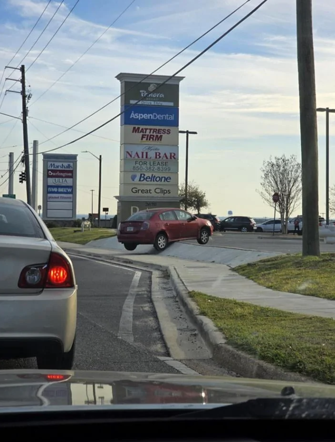 Červené auto zaparkované na obrubníku vedľa obchodného centra a značka s názvami obchodov v pozadí.
