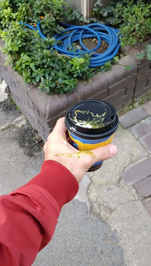 Muž v červenej bunde drží rozliaty nápoj so zelenou škvrnou na viečku, chodník v pozadí.