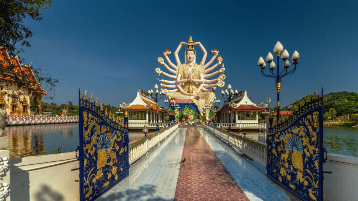 Socha s mnohými rukami v chráme na pozadí modrej oblohy a zdobenej brány.