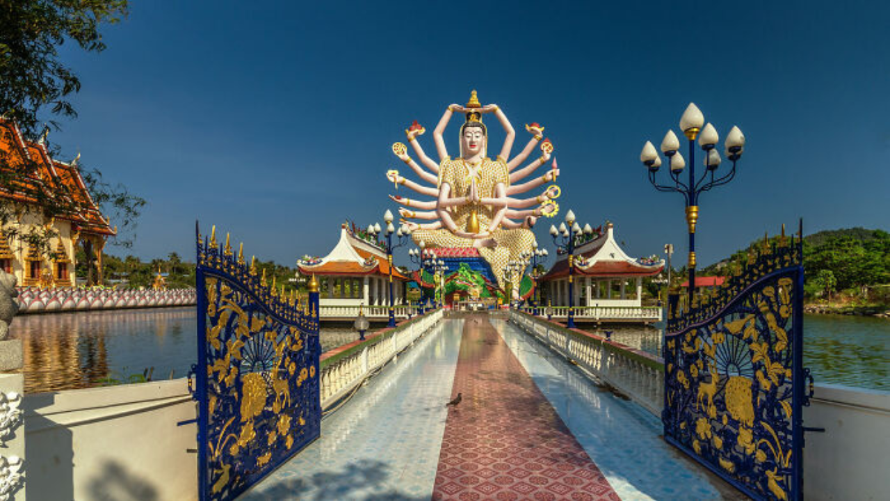 Socha s mnohými rukami v chráme na pozadí modrej oblohy a zdobenej brány.