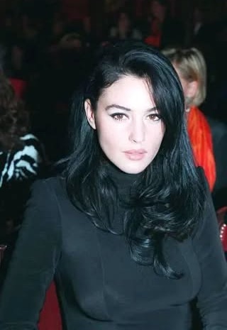 Osoba s čiernymi vlasmi v elegantnom čiernom oblečení sedí v interiéri, za ňou rozmazaní ľudia.