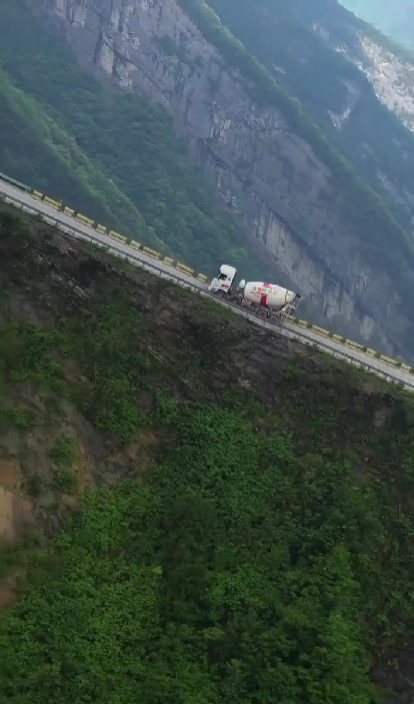 Cementový mixér na úzkej horskej ceste, obklopený strmými svahmi a zelenými úbočiami.