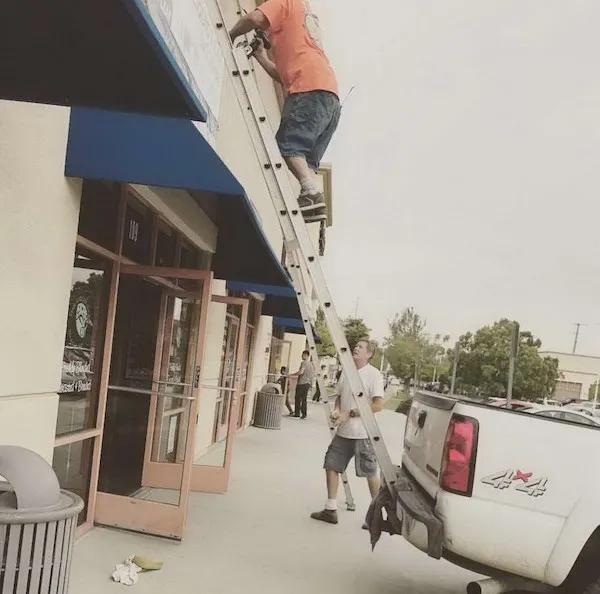 Muž na rebríku opretom o auto vykonáva prácu na budove, zatiaľ čo druhý muž ho podopiera.