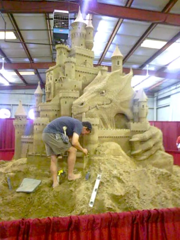Majster tvaruje pieskový hrad s dračou sochou na výstave v hale, jemné detaily prilákajú obdiv.