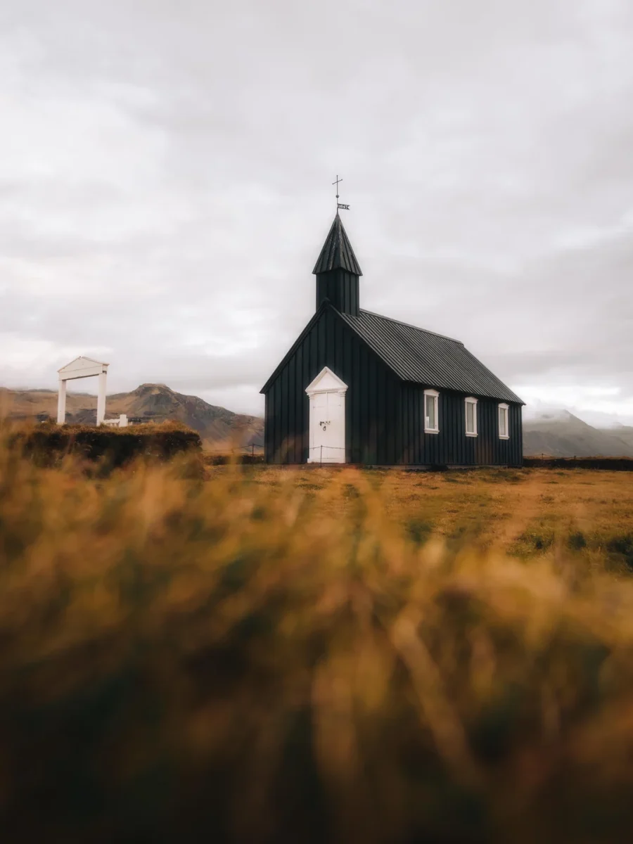 Malý čierny kostol na vidieku obklopený trávnatými kopcami pod zamračenou oblohou.