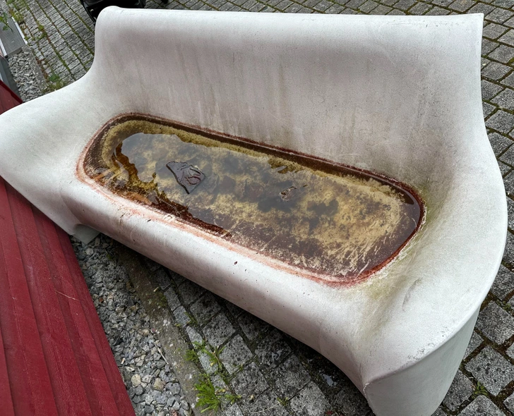 Betónová lavička zadržiavajúca dažďovú vodu na povrchu, nachádza sa na dlažbe vedľa červenej štruktúry.