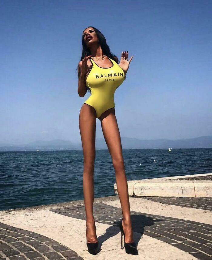 Osoba v žltých plavkách na móle pri mori s jasnou oblohou a horami v diaľke.