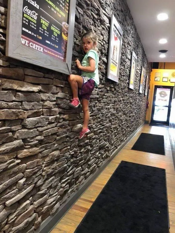 Dieťa lezie po kamennej stene v interiéri, ku ktorej je pripojené menu reštaurácie.