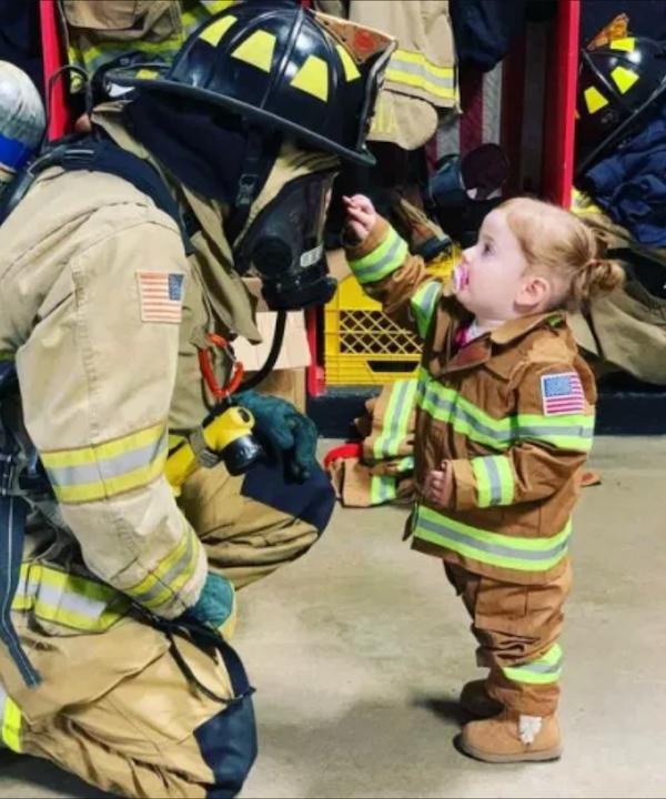 Dieťa v hasičskom oblečení pomocou bábätka skúma prilbu skutočného hasiča, vnútorný priestor stanice.