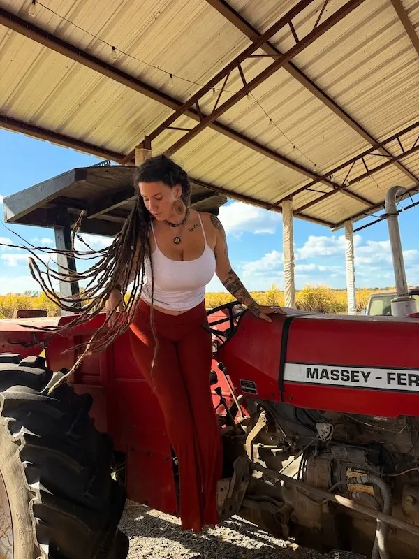 Žena s dlhými dredmi stojí na červenom traktore pod zastrešeným prístreškom za slnečného dňa.
