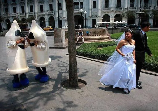 Nevesta a ženích kráčajú po námestí, sleduje ich dvojica v kostýmoch kondómov, fontána v pozadí.