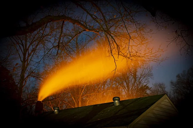 Oranžový dym vychádzajúci z komína domu na tmavej oblohe, obklopený stromami bez lístia.