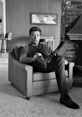 Muž sediaci v kresle, obutý v topánkach, interiér s krbom a obrazom na stene.