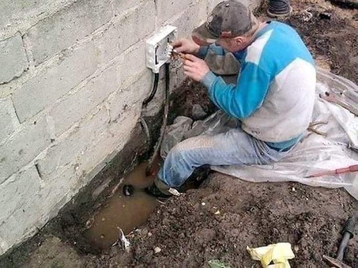 Osoba opravuje vonkajšiu elektrickú zásuvku v zemi, sedí blízko steny nad blatistou vodou.