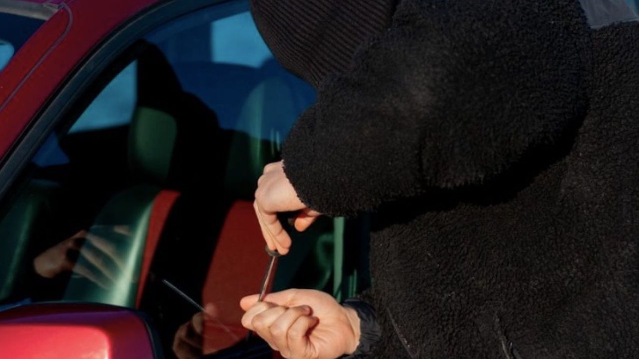 Osoba v čiernom oblečení pokúšajúca sa vlámať do červeného auta s nástrojom v ruke.