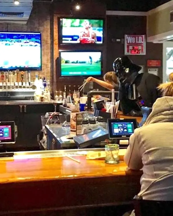 Osoba v kostýme obsluhuje bar, zatiaľ čo na TV obrazovkách bežia športové programy.