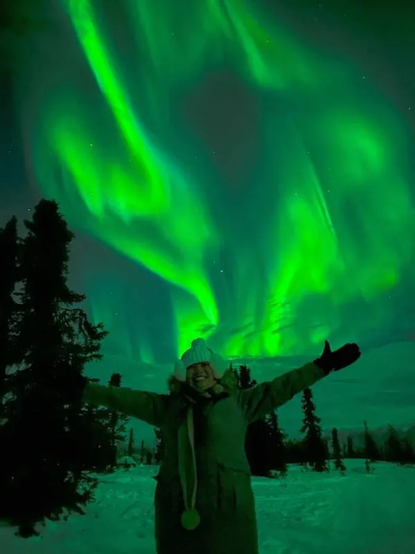 Osoba na zasneženom poli pod žiarivou zelenou polárnou žiarou, obklopená tmavými stromami.