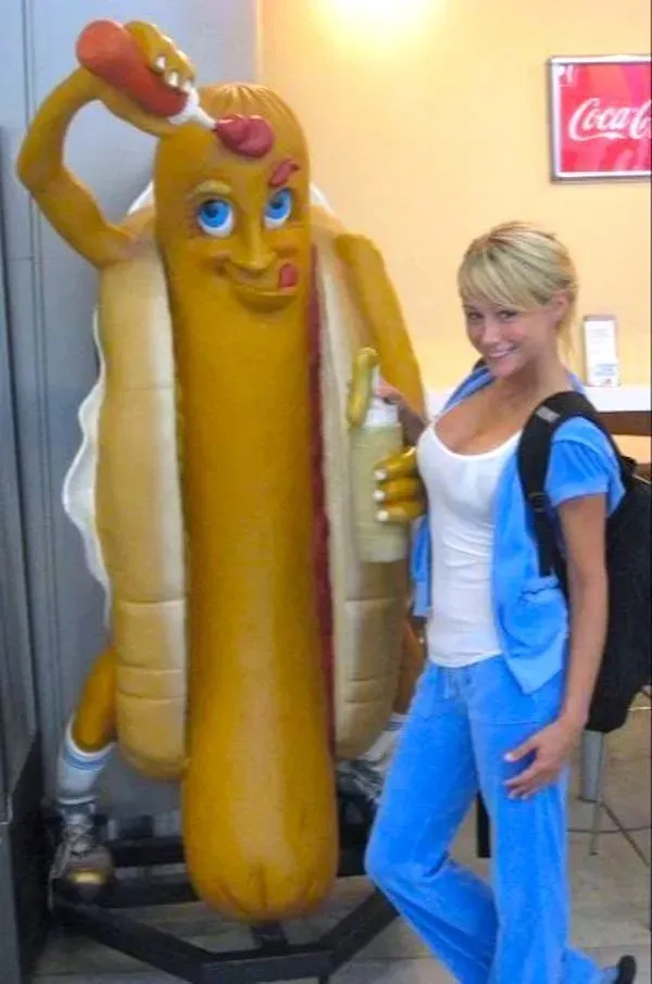 Žena v modrom pózuje vedľa sochy hotdogu s kečupom a horčicou.