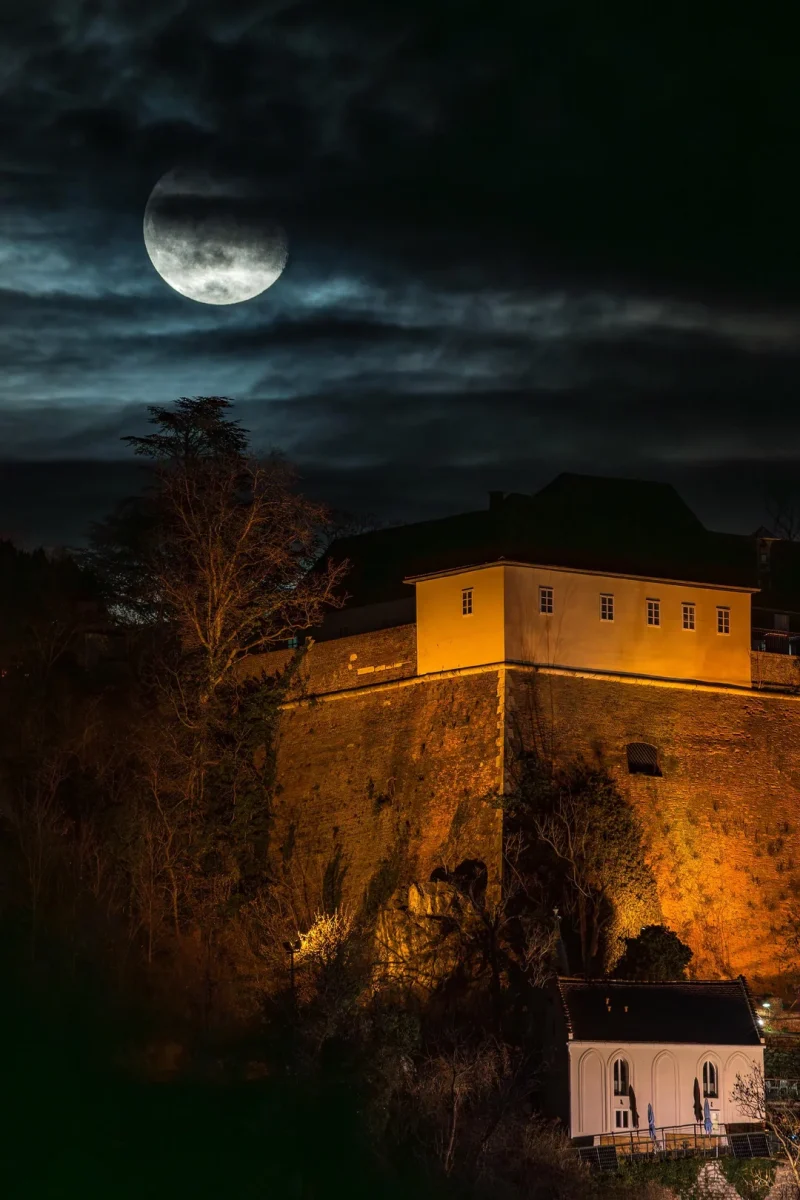 Mesačný svit nad osvetlenou hradbou a stromami počas tajomnej noci.
