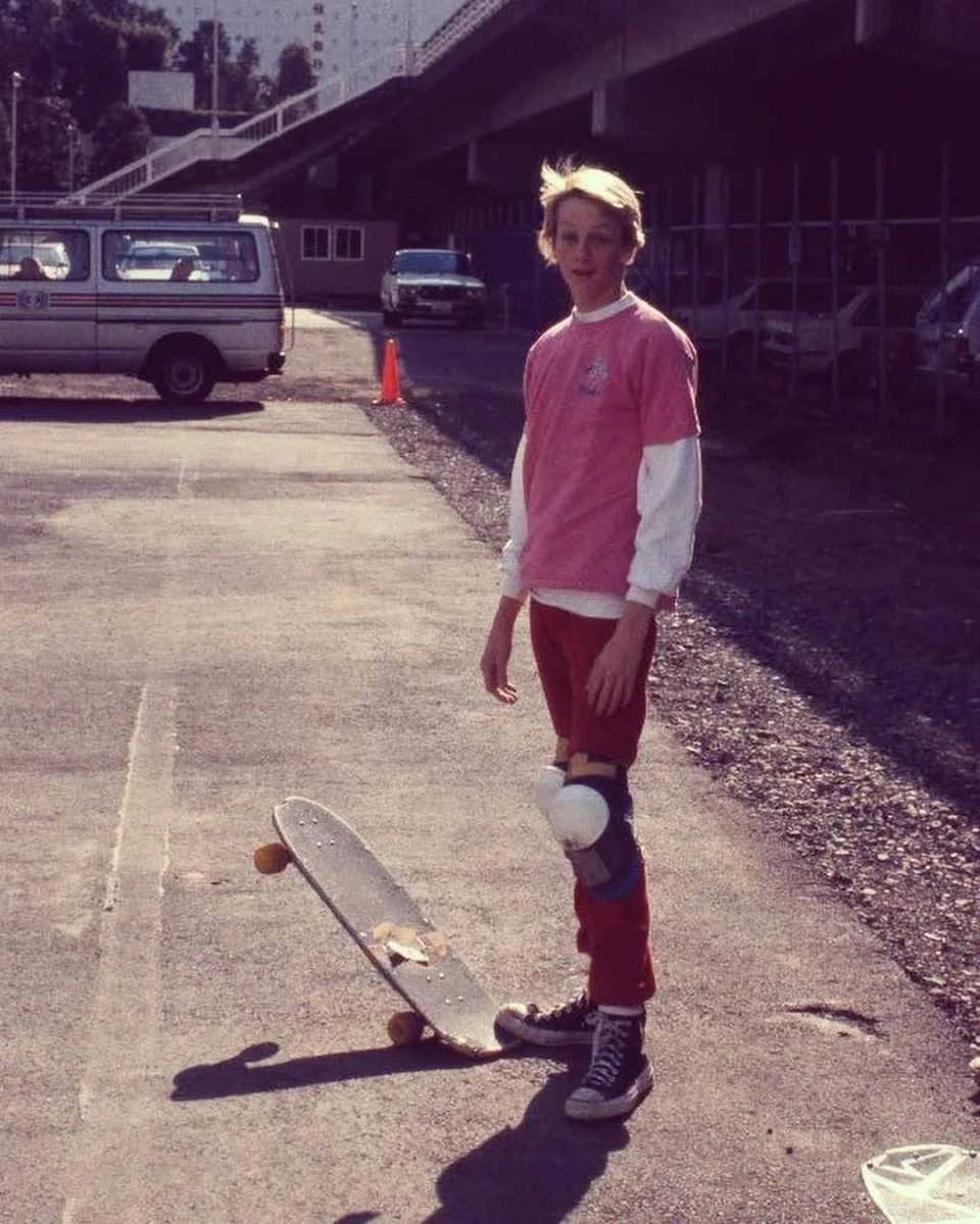 Osoba na skateboarde s chráničmi kolien v ružovom tričku na mestskom chodníku.