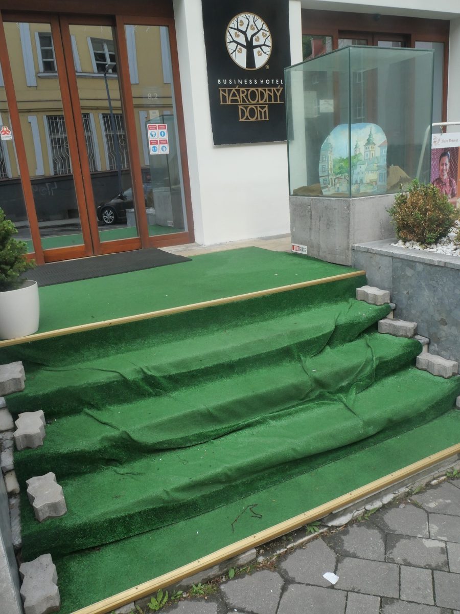 Vchod do hotela s nápisom Národný dom, zelené schody s umelou trávou a vitrína s obrazom.