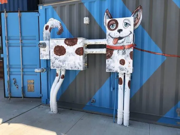 Umelecká inštalácia v tvare psa z potrubia a kovových krabíc na modrom pozadí kontajnera.