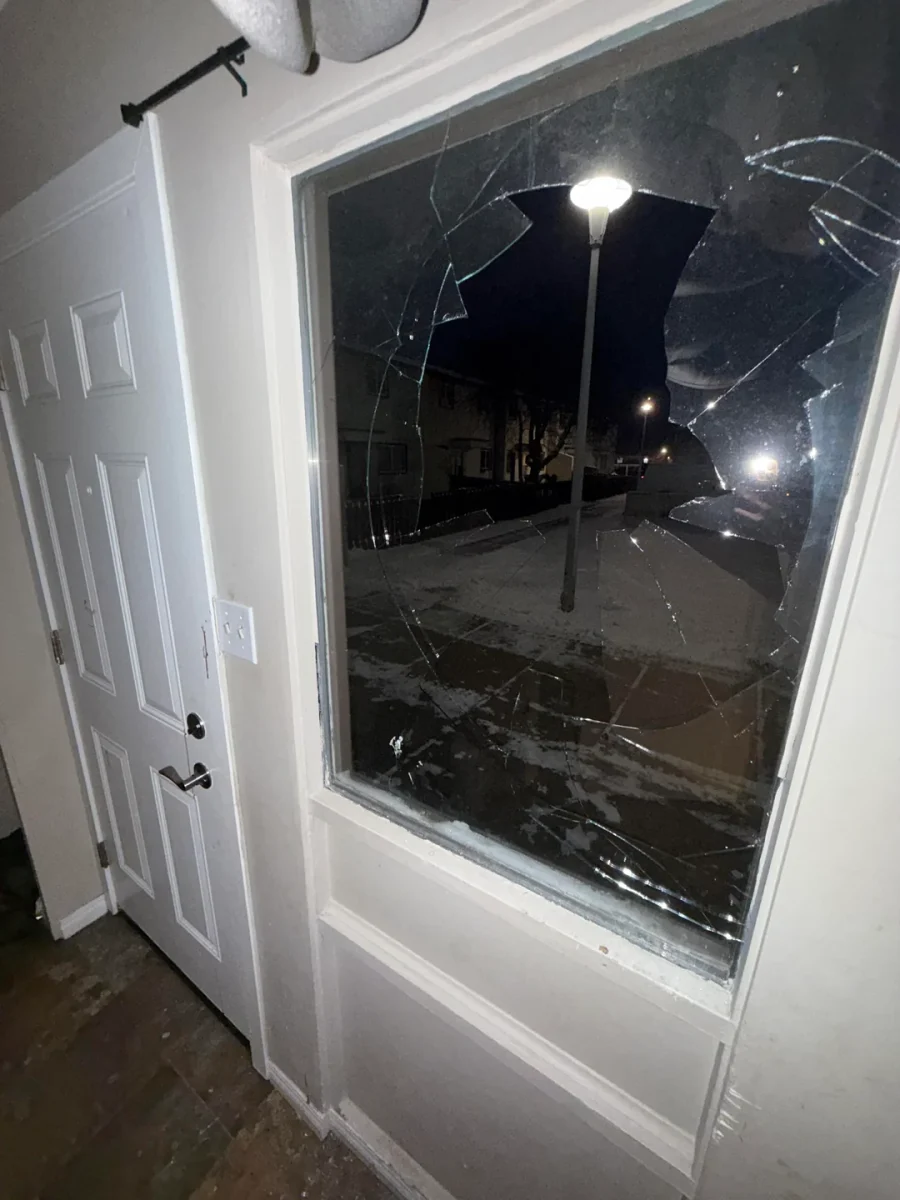 Rozbité okno vo dverách, nočný pohľad na osvetlenú ulicu, lámané a prasknuté sklo zblízka.