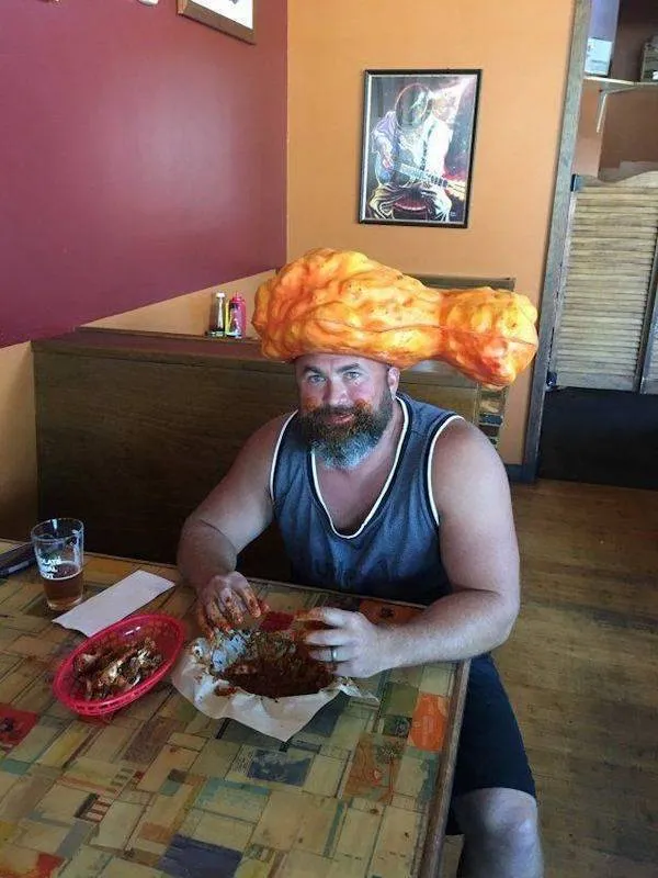 Osoba s klobúkom v tvare kuracieho stehna sedí v reštaurácii, stoluje s úsmevom.