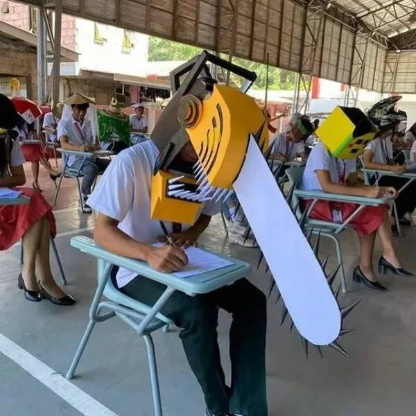 Študent so strojovou maskou píše test, zatiaľ čo ostatní v triede majú rôzne kreatívne pokrývky hlavy.