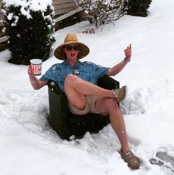 Osoba v letnom oblečení s klobúkom sedí na kresle v snehu, s nápojom v ruke a užíva si zimu.