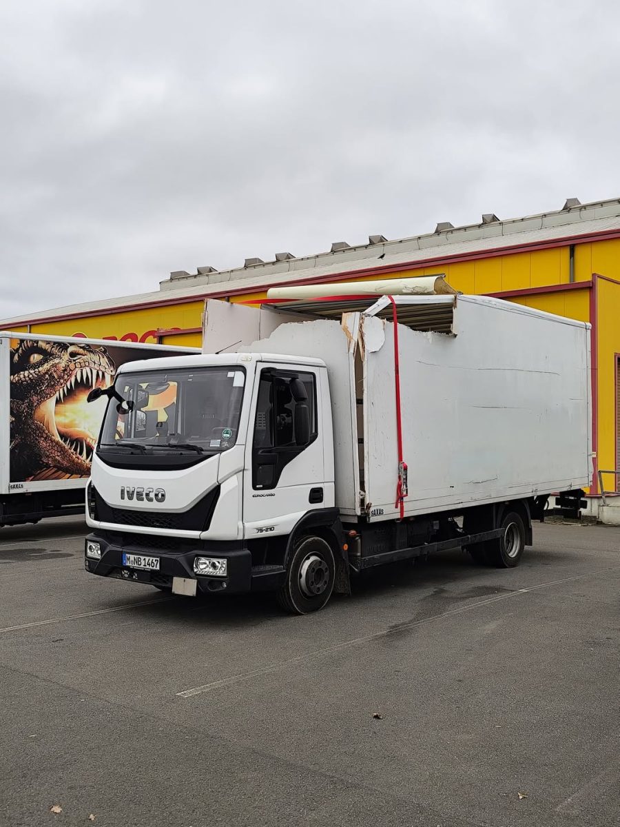 Biely nákladný automobil s poškodenou plošinou stojí na parkovisku pred žlto-červenou budovou.