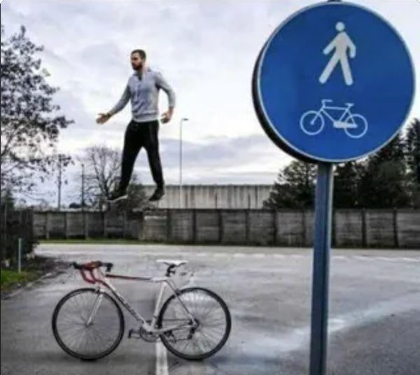 Osoba akoby levituje vedľa cykloznačky a bicykla na ceste, optická ilúzia v mestskom prostredí.