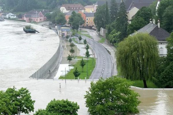 Povodeň pri rieke zaplavuje cestu v meste, znemožňuje dopravu a zasahuje do mestskej zelene a budov.
