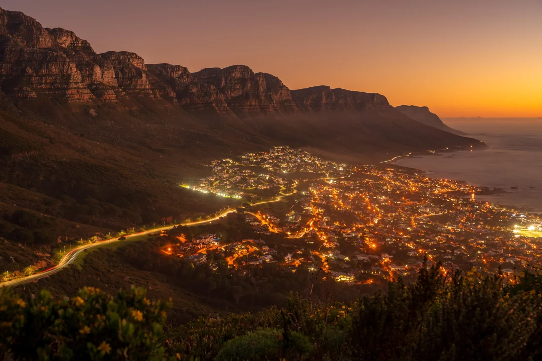 Nočná panoráma mesta pri pobreží s osvetlenými ulicami a horami v pozadí.