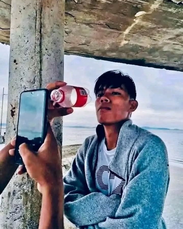 Ľudia s mobilom fotia muža pijúceho z fľaše pri pláži, opretého o betónový stĺp pod baldachýnom.