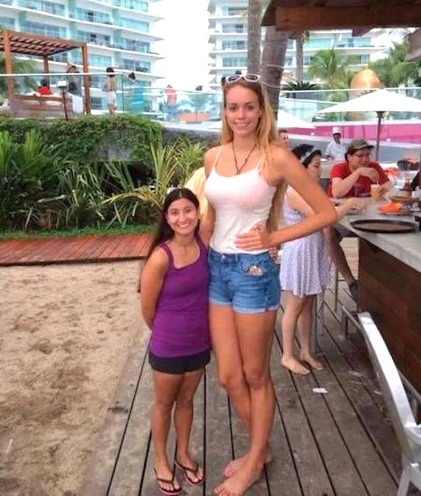 Dve ženy stojace vedľa seba na pláži so zreteľným výškovým rozdielom, slnečný deň.