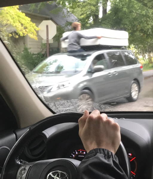 Osoba na streche auta drží matrac počas jazdy. Pohľad z auta na scéne počas daždivého dňa.