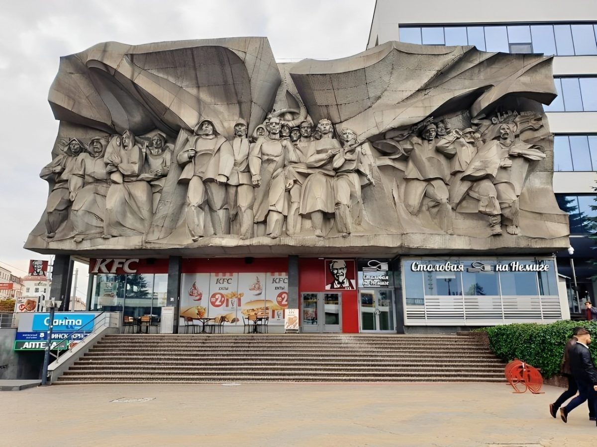 Pamätný reliéf nad reštauráciou KFC pri obchodnom centre; schody a dve osoby prechádzajúce popred budovu.