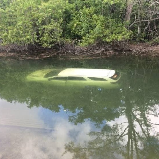 Auto pod vodou blízko brehu, zrkadliace sa stromy a obloha na hladine v prírodnom prostredí.