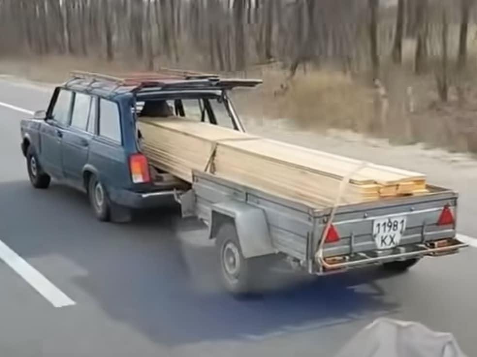 Auto s prívesom preváža dlhé drevené dosky na diaľnici,ťahané starším modelom vozidla.