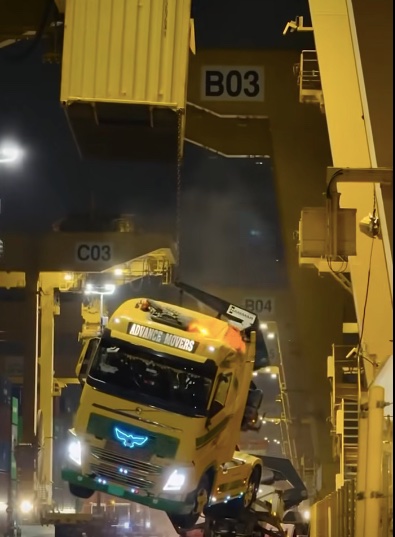 Nákladný žeriav v prístave manipuluje s kontajnerom nad kamiónom pri nočnej obsluhe.