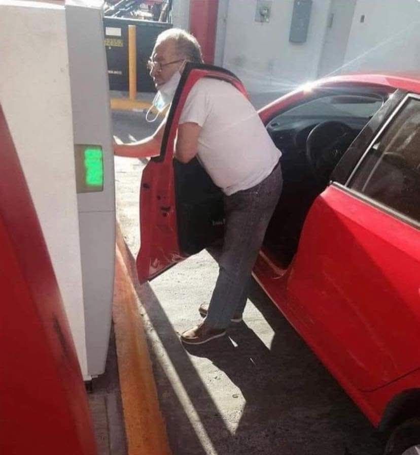 Muž pri červenom aute využíva platobný automat so stiahnutou rúškou pod bradou na parkovisku počas slnečného dňa.