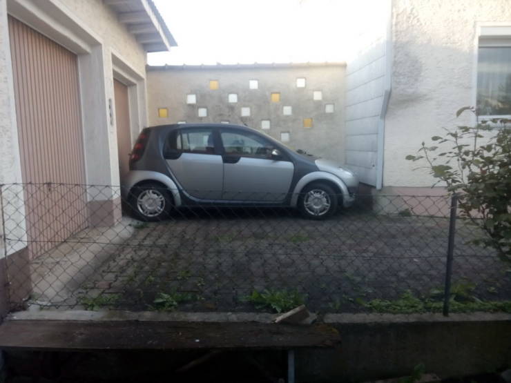 Malé strieborné auto zaparkované na úzkom dvore medzi domom a garážou.